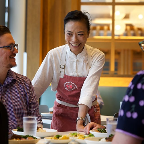 Person in a pink apron serving food to diners at a restaurant table.
