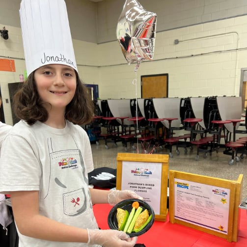 Kid in a chef's hat and gloves presents a plate of food in front of a display table with a red tablecloth, which holds framed documents and a silver balloon, suggesting a cooking event or competition.