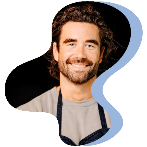 Person wearing a light gray shirt and a dark apron stands against a black background with a blue abstract shape framing the image.