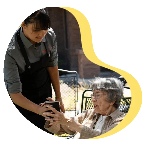 Person in uniform gives a cup to seated elderly person outdoors, framed by a yellow abstract shape.