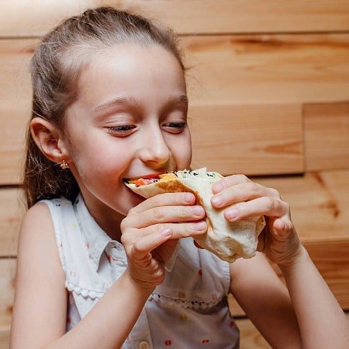 A young student smiling while eating a wrap-style meal, representing healthy and enjoyable school dining.