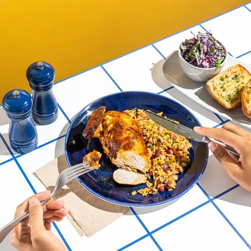 Close-up of a grilled chicken breast on a blue plate served with seasoned rice and beans, while hands use a fork and knife to cut the food on a tiled table setting.