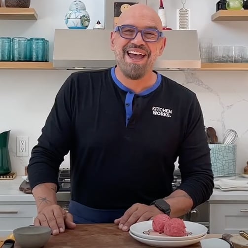 A bald man in glasses smiles while holding a plate of meatballs, showcasing a joyful moment.