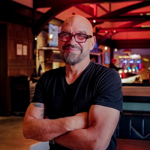 Person standing with crossed arms in black t-shirt inside dimly lit restaurant with tables and chairs.