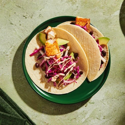 Two tacos with grilled tofu, avocado, red cabbage, black beans and sauce on a green plate.