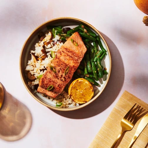 A plate with grilled salmon, green beans, rice with mushrooms, and a grilled lemon slice, next to a gold fork and knife.
