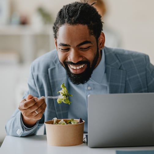 Person in a blue suit eating a salad while working on a laptop.