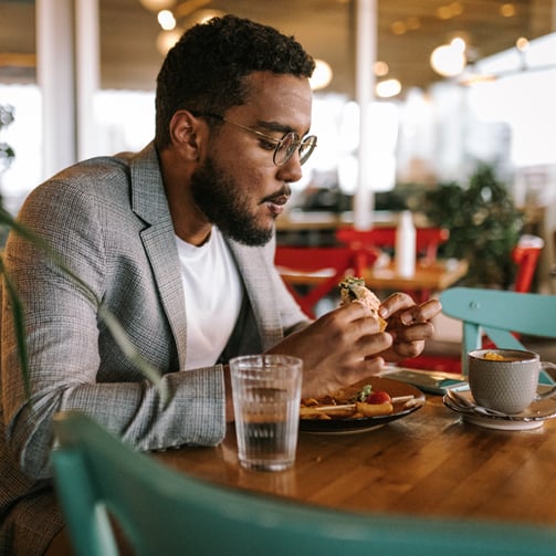 Person in gray blazer eating a sandwich at a wooden table with water glass, bowl, and plate of food in a restaurant.
