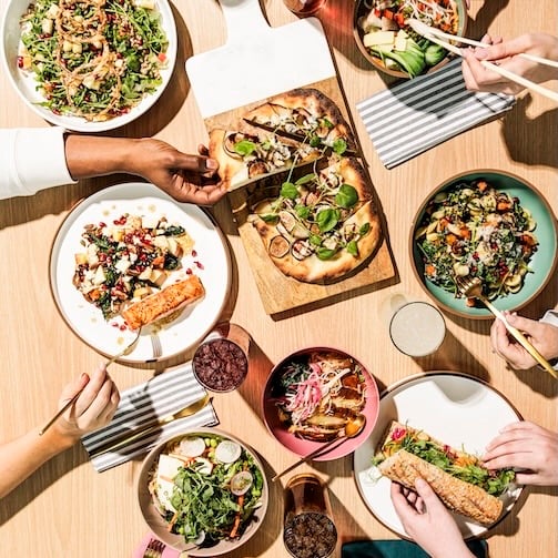Overhead view of a shared meal with pizza, salads, wraps, and hands reaching across a wooden table.