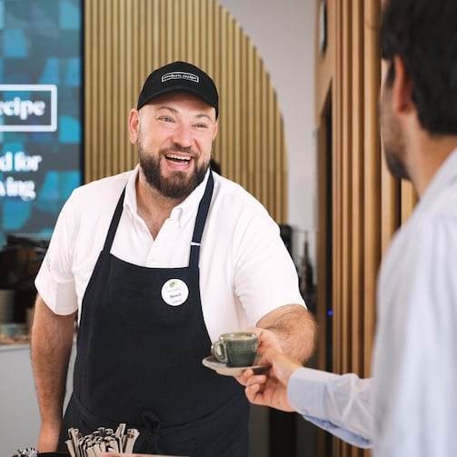 Cafe staff member in an apron handing a cup of coffee to a customer across the counter in a modern café.
