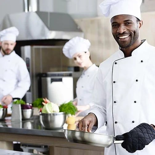 Chefs in white uniforms and hats prepare food in a professional kitchen; one chef holds a frying pan wearing an oven mitt.