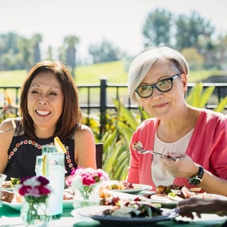 Two older adults sit outdoors at a sunlit table enjoying a meal together, surrounded by greenery and flowers.
