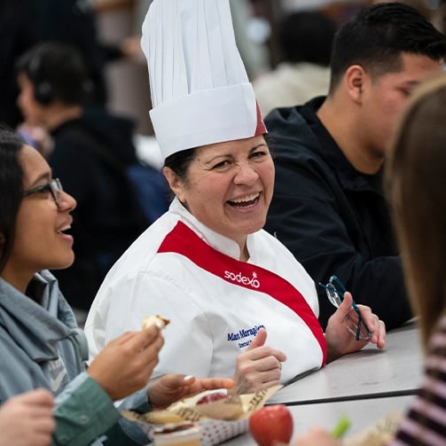 A smiling woman in a chef hat enjoys a plate of food, showcasing her passion for cooking and culinary delight.