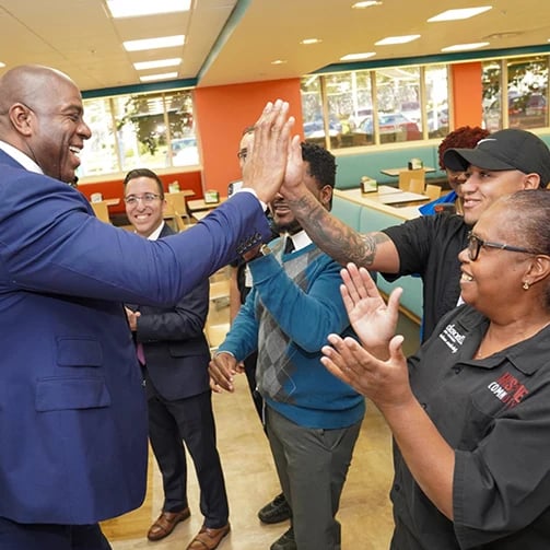 Five people standing around tables, exchanging high-fives and clapping in a bright room.