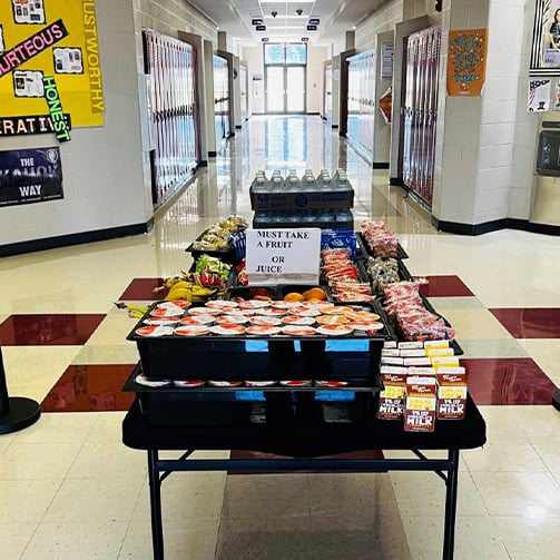 Mobile breakfast cart stocked with packaged meals milk juice and fruit in a school hallway, featuring a sign that reads Must Take a Fruit or Juice, with lockers lining both sides of the corridor.