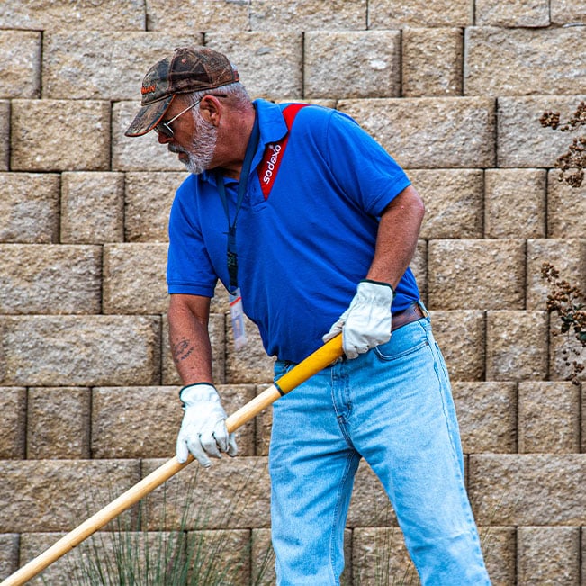Facilities maintenance worker using a long-handled landscaping tool beside a stone retaining wall, performing outdoor groundskeeping and property upkeep in a commercial or campus setting.