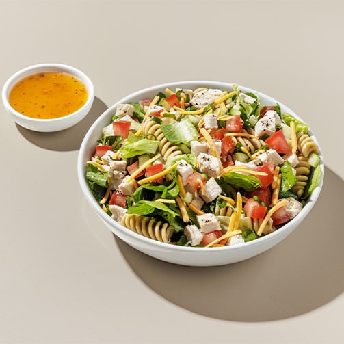 Bowl of pasta salad with lettuce, tomatoes, cheese, chicken, and dressing on the side.