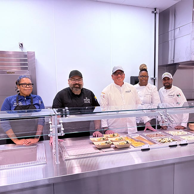 School food service team standing behind a cafeteria serving counter with prepared meals on display, highlighting culinary operations, meal preparation, and student dining support in an educational setting.
