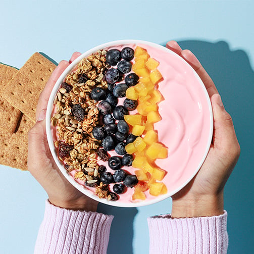 Hands holding a pink smoothie bowl topped with granola, blueberries, and diced fruit, with graham crackers on the side, showcasing a colorful and nutritious breakfast or snack option.