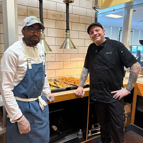 Two culinary staff members standing beside a school cafeteria hot food station, highlighting food service operations, meal preparation, and nutritious dining programs in an educational setting.