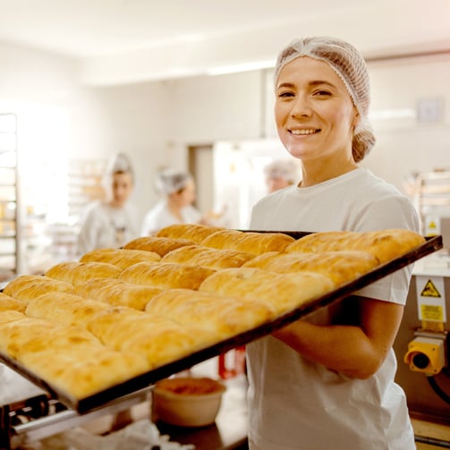 A smiling baker wearing a hairnet holds a large tray of golden pastries in a busy bakery