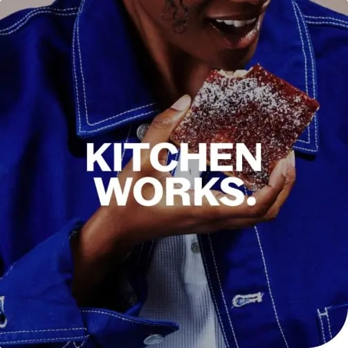 Person in a blue jacket holds a powdered sugar-covered brownie or cake; white text across the center reads KITCHEN WORKS.