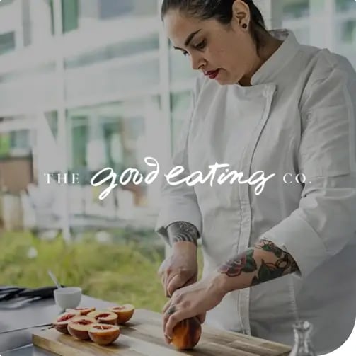 A woman in a chef's uniform slices an orange on a cutting board in a bright kitchen.