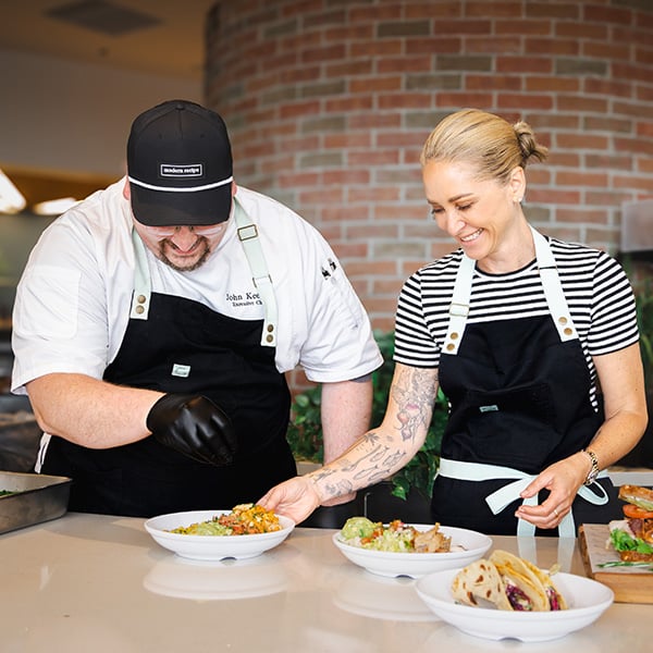 Two chefs wearing black aprons plating dishes on a counter in a modern kitchen, with bowls of prepared food and tacos arranged nearby against a brick wall background.