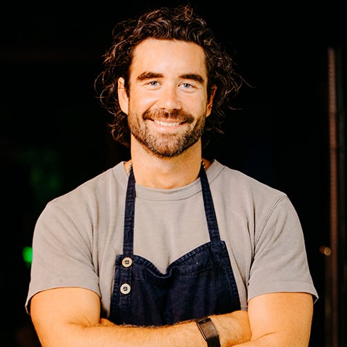 Portrait of a Gabe Kennedy wearing a T-shirt and dark apron, standing with arms crossed against a dark background in a kitchen or restaurant setting.