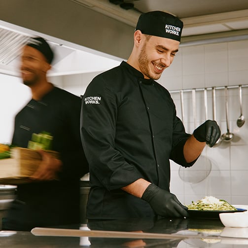 A chef seasoning a dish while other workers are in motion in a busy kitchen