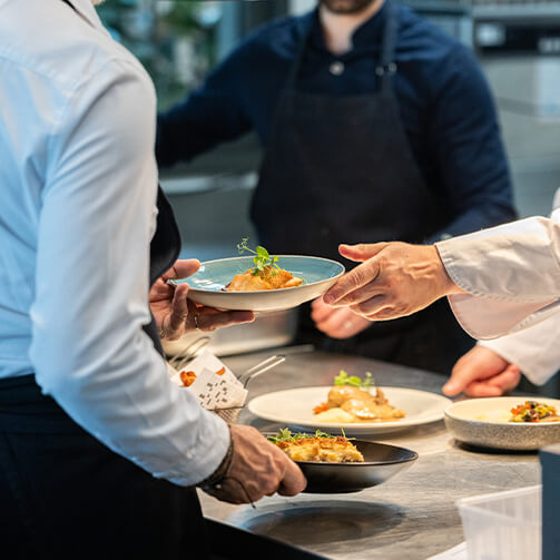 Image depicting a professional kitchen environment where chefs are collaboratively plating and preparing dishes. One chef is handing a plate of food to another, while multiple dishes are visible on the counter, showcasing the teamwork and coordination of culinary professionals