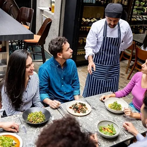 Group of people sit around a table at a restaurant with plates of food, while a chef or waiter in a striped apron speaks to them; wine bottles are visible in the background.