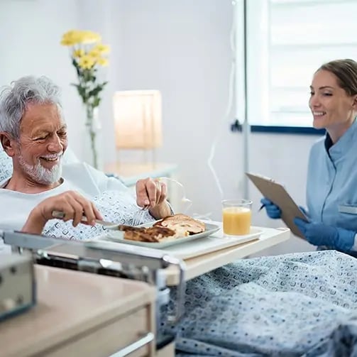 Elderly person eats a meal from a tray in a hospital bed while a healthcare worker with a clipboard sits nearby; flowers and a lamp are visible in the background.