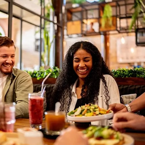 Server places a salad in front of a person with long curly hair seated at a wooden table in a cozy restaurant with plants, warm lighting, and visible food and drinks.