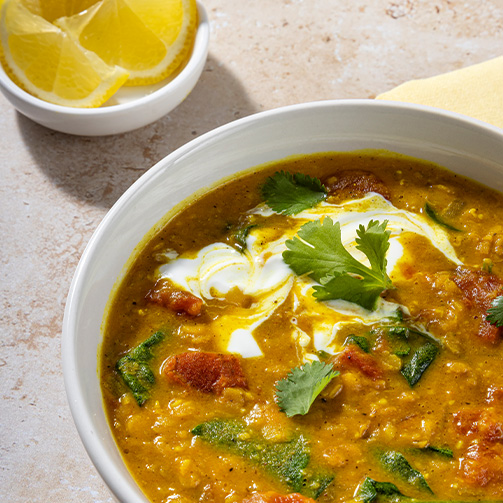 Bowl of creamy vegetable soup topped with yogurt, fresh cilantro, leafy greens, and crispy croutons, served alongside lemon slices, highlighting a wholesome and flavorful meal option.