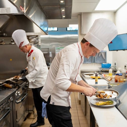 Two chefs in white uniforms working together to prepare meals in a senior living community kitchen.