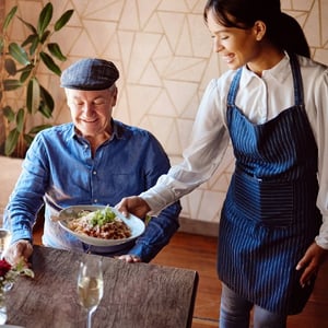 A smiling server delivers a dish to a senior man seated at a table in a modern dining space.