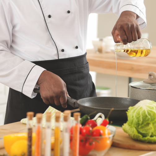 Image of a chef in a white jacket and black apron pouring oil into a frying pan in a busy kitchen with fresh ingredients.