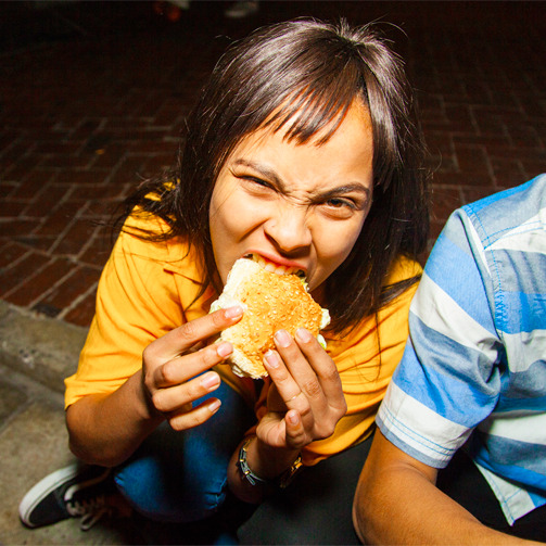 A female college student taking a bite of a burger while posing for the camera.