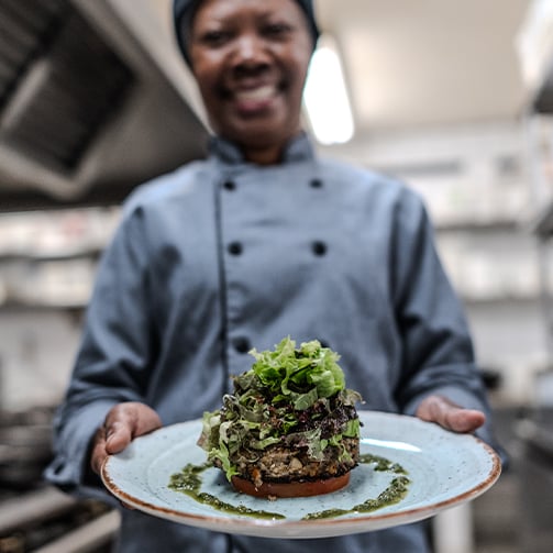 Chef in a professional kitchen holds a plate with a gourmet dish topped with fresh greens and garnished with green sauce on a rustic base.