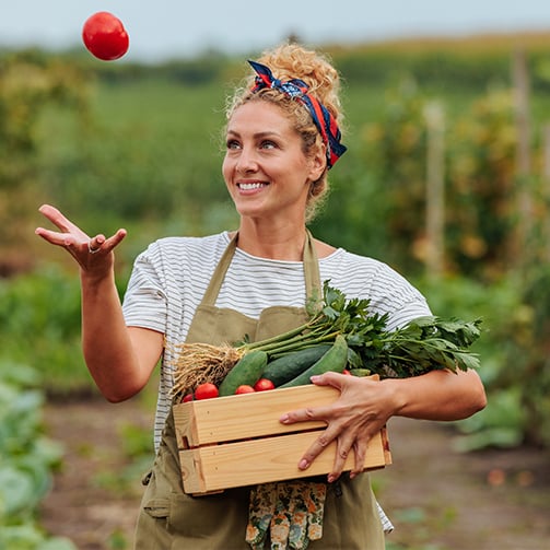 Person in apron holding a crate of vegetables and tossing a tomato in a garden with green plants in the background.