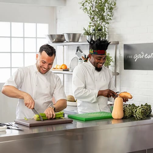 Two chefs chopping celery and peeling squash in a modern stainless-steel kitchen.