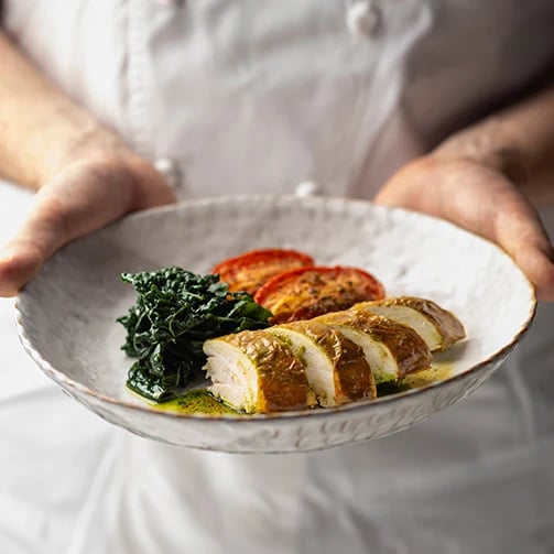 Chef holding plate of sliced chicken breast over sauteed greens with roasted tomatoes
