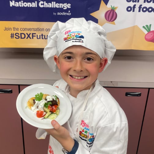 Child wearing a white chef's coat and hat holds a plate of food with cherry tomatoes, cucumber slices, white sauce, and parsley garnish, standing in front of a 'National Challenge by Sodexo' banner