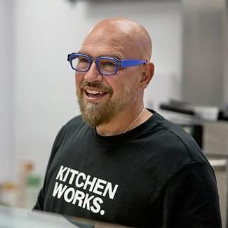 Chef Michael Symon wearing blue-framed glasses and a black “KITCHEN WORKS.” T-shirt in an indoor setting.