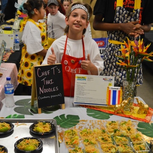 Child at food fair table giving thumbs up behind trays of Thai noodles, with recipe sign and classmates.