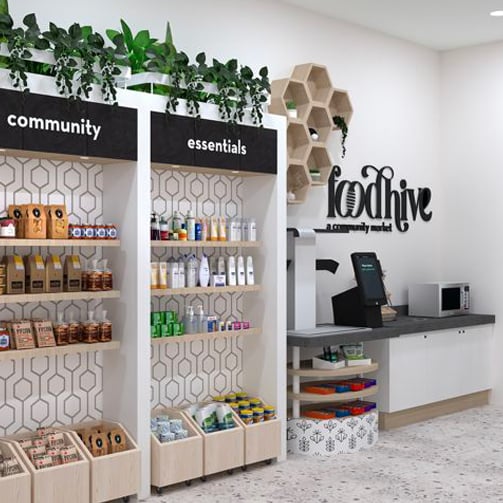 Modern self-serve community pantry with shelves labeled community and essentials, stocked with food and personal care items, plants on top, and a counter with a kiosk under a food hive sign.