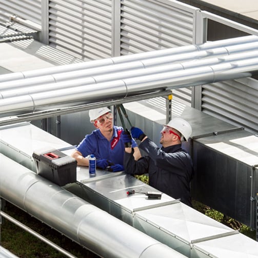 Two workers wearing hard hats and safety gear performing maintenance on large industrial piping and HVAC equipment on a rooftop facility, surrounded by metal ducts and utility infrastructure.