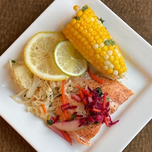 Close-up of a plated meal featuring baked salmon topped with shredded vegetables, served with corn on the cob, lemon and lime slices, and a side of pickled vegetables on a square white plate.