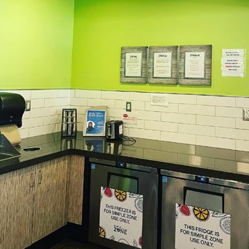Break room or food storage area with green walls, countertop appliances, framed notices, and two undercounter refrigerators labeled as dedicated “Simple Zone” storage for specific food items.